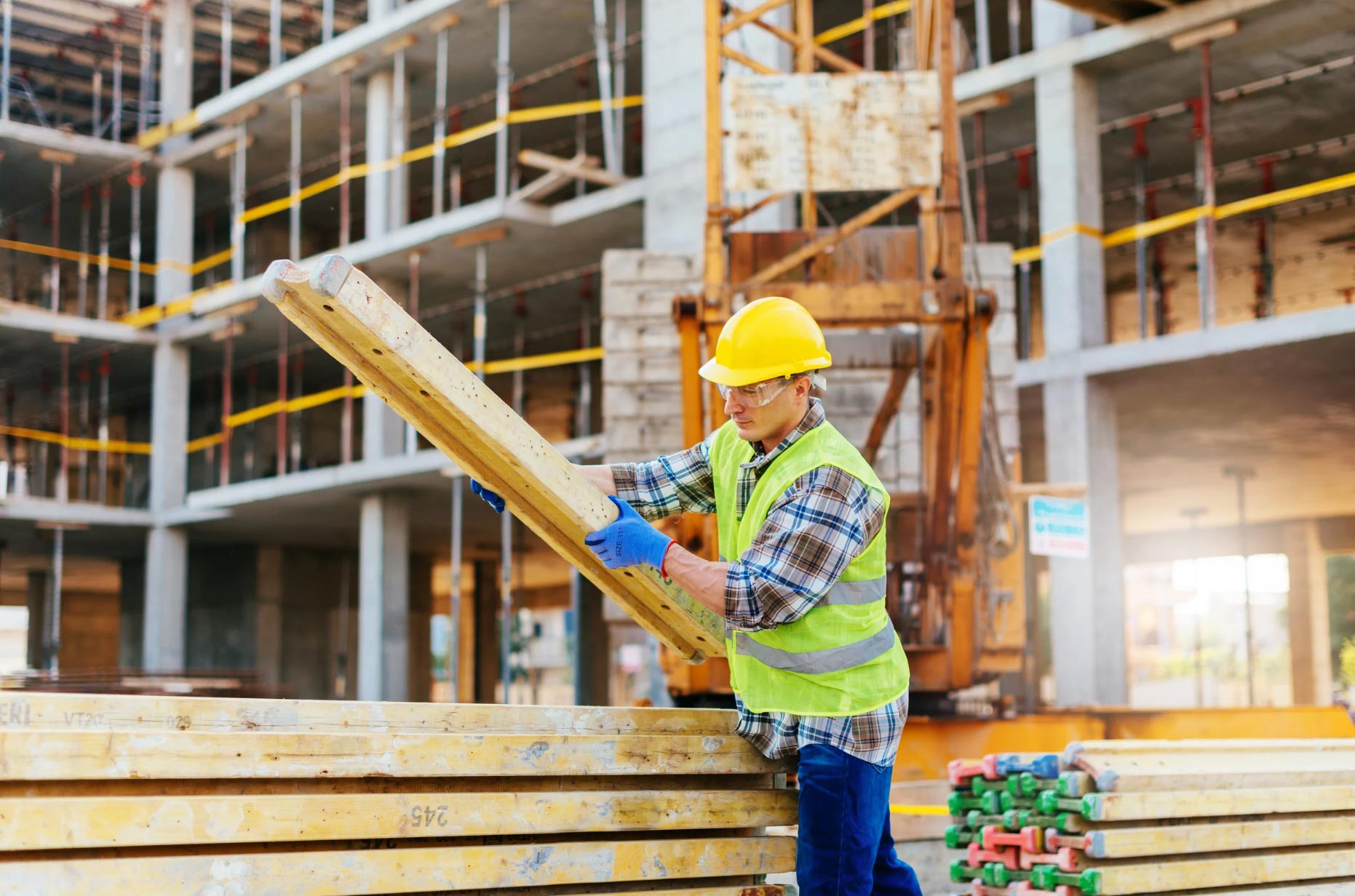 A construction worker in safety gear expertly handles wooden planks at a bustling Nextt Government Construction site, with a building frame rising proudly in the background.